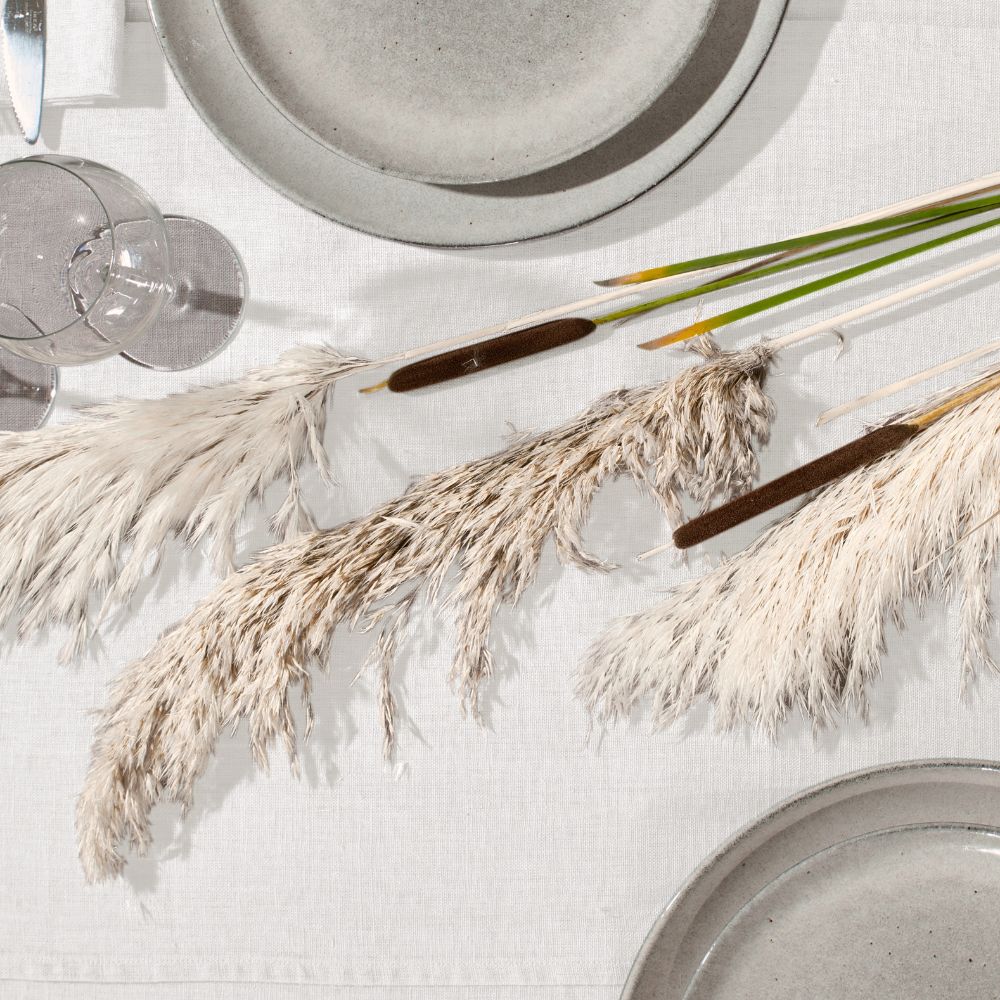 Gedeckter Tisch von oben mit weißer Leinen-Tischdecke, Steingut-Tellern, Weingläsern und Pampasgras